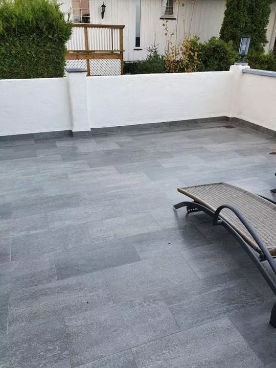 Romslig terrasse med grå fliser og en hvit vegg, med en lenestol i kurvmateriale på gulvet.