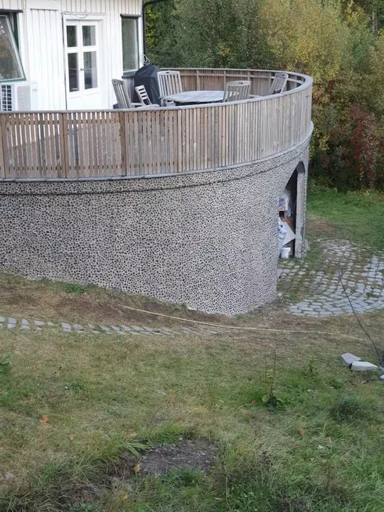 Rund terrasse med trebalkong og dekorert murvegg, sett fra en hage.