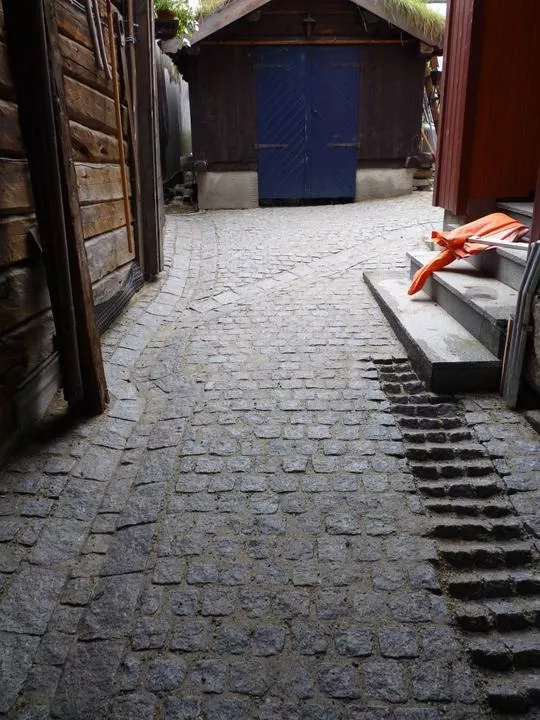 Detaljert brosteinlagt bakgård med grå steinheller, omgitt av gamle trebygninger og en trapp i bakgrunnen.